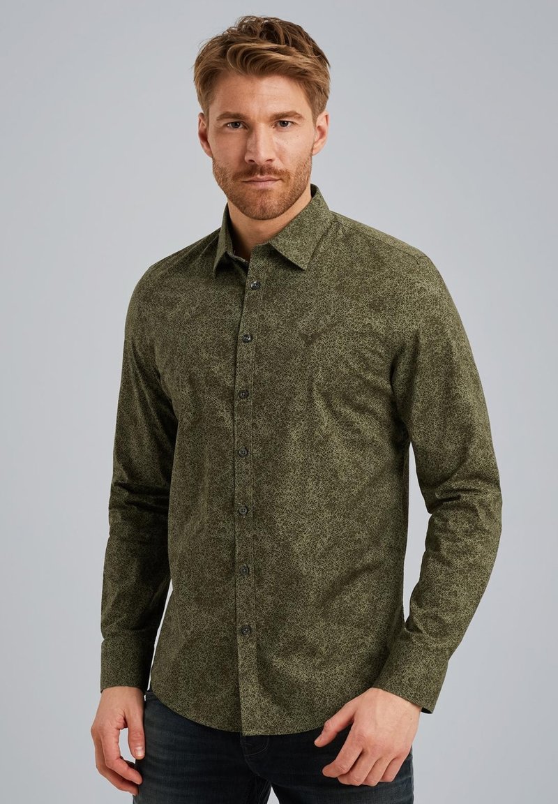 Chemise à manches longues verte à motif, avec un col à boutons, présentant une surface texturée et des boutons noirs, portée sur un jean noir.