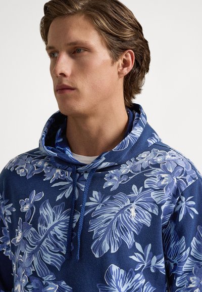 Jovem com cabelo castanho claro a usar um pólo com capuz azul com padrões de folhas tropicais e flores em azul claro e branco, virado para a esquerda.