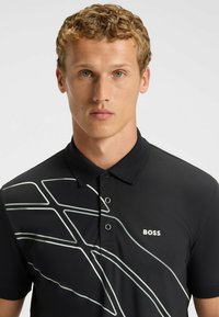 Czarna koszulka polo z kołnierzykiem, ozdobiona kontrastującymi białymi wzorami w linie. Z przodu znajdują się trzy guziki oraz białe logo.