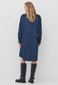 Traper haljina s dugim rukavima, tamnoplave boje, s teksturiranim crnim detaljima na ramenima; uparena s čizmama od crne kože do koljena.