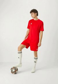 Nuori mies punaisessa jalkapallosarjassa ja valkoisissa kengissä lepuuttaa jalkaansa mustavalkoisen jalkapallon päällä yksinkertaista valkoista taustaa vasten.