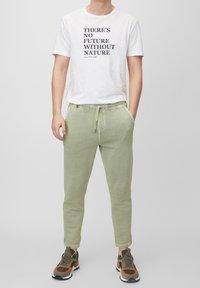 Vit t-shirt med svart text som säger "DET FINNS INGEN FRAMTID UTAN NATUREN." Ljusgröna joggerbyxor med dragsko och fickor, tillsammans med sneakers.