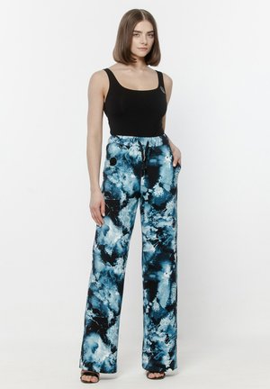 Kobieta w czarnym, bezrękawnym topie i szerokich, niebiesko-białych spodniach tie-dye, stojąca z jedną ręką w kieszeni na białym tle.