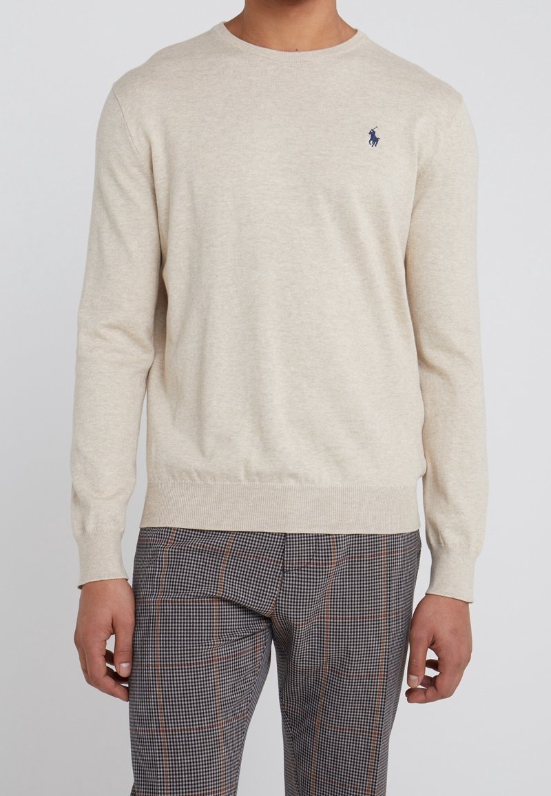 Homme portant un pull beige clair à manches longues avec un petit logo sombre sur la poitrine et un pantalon à carreaux avec un motif marron et noir.