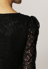 Top de encaje negro con un diseño floral texturizado, hombros abullonados y mangas largas transparentes. El escote es redondeado y elegante.