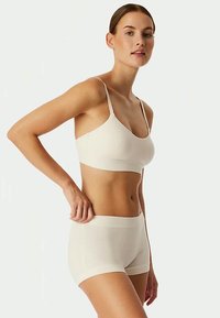 Kvinde justerer en sømløs lys beige bh og matchende boyshort underbukser mod hvid baggrund.