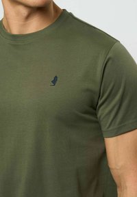 Vīrietis valkā olīvu zaļas krāsas T-kreklu ar apaļu kakla izgriezumu un nelielu tumšu izšūtu logotipu kreisajā krūtīs, redzams no kakla līdz viduklim.