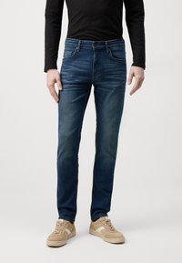 Jeans en denim bleu à coupe skinny avec un effet délavé, un design à cinq poches et une fermeture bouton, associés à des baskets de couleur claire.