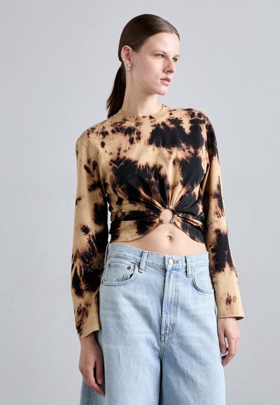 Dlouhý rukáv, tie-dye top v béžové barvě s černými vzory, se zavázaným předním dílem a dvěma dekorativními kovovými kroužky; spojen s světle modrými džínami.
