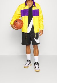 Persona sosteniendo un balón de baloncesto, vistiendo una chaqueta amarilla y morada de los Lakers, pantalones cortos negros y blancos de Nike, calcetines blancos y zapatillas negras y blancas.