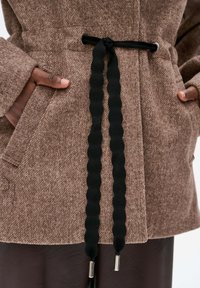 Bruin gestructureerde jas met een zwarte elastische trekkoordsluiting. Bevat zakken en zilveren hardwareaccenten. Gezellig, warme stof.