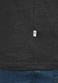 T-shirt gris foncé en tissu doux, avec une petite étiquette blanche comportant un point d'exclamation sur la couture latérale, portée rentrée dans un jean bleu.