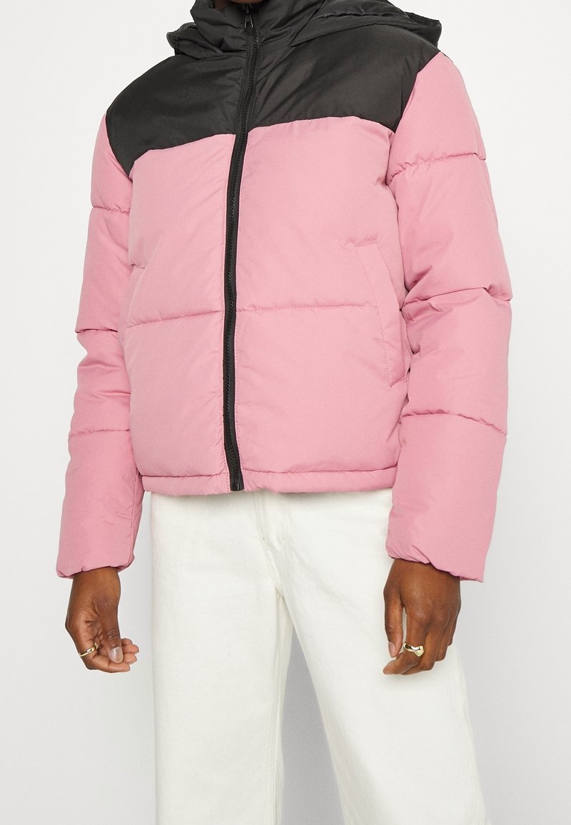 Giacca a vento con una sezione inferiore rosa e una superiore nera, dotata di chiusura con zip e due tasche laterali. Materiale sintetico e liscio.