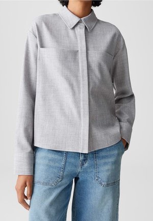 Personne portant une chemise gris clair à manches longues boutonnée avec col et un jean bleu, debout avec une main dans la poche.