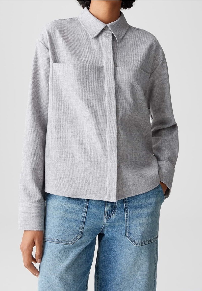 Personne portant une chemise gris clair à manches longues boutonnée avec col et un jean bleu, debout avec une main dans la poche.