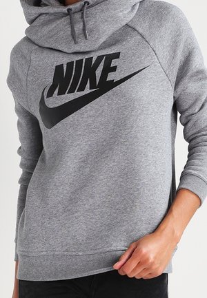 Personne portant un sweat à capuche gris Nike avec un grand logo noir, une main tenant l'ourlet inférieur, pantalon noir visible.