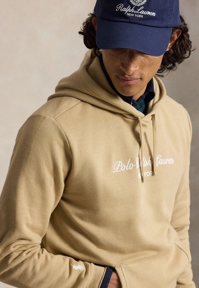 Νεαρό άτομο φοράει ένα μπεζ φούτερ Polo Ralph Lauren και μια σκούρα μπλε καπέλο Ralph Lauren, κοιτάζει προς τα κάτω με τα χέρια στις τσέπες του φούτερ.