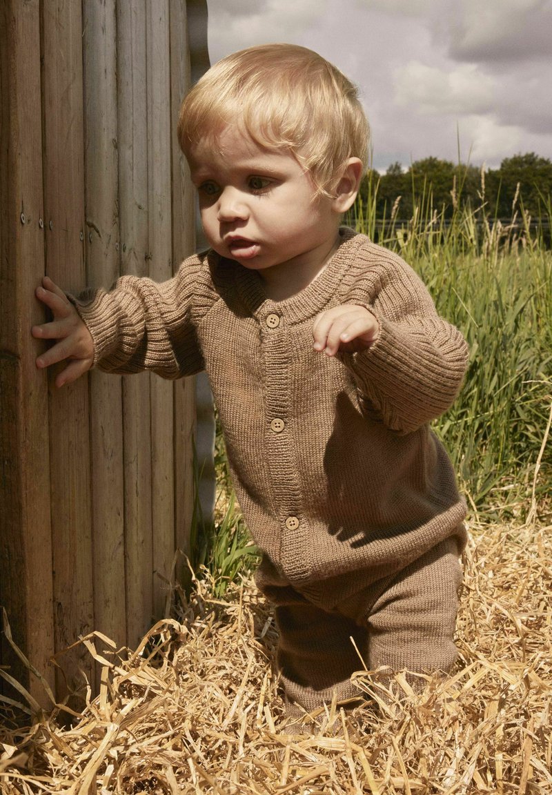 Tout-petit blond portant une tenue en tricot marron, debout sur de la paille, tendant la main pour toucher une clôture en bois dans un espace herbeux extérieur.