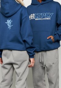 I felpe con cappuccio blu presentano grandi grafiche frontali e un logo più piccolo sul retro. Abbinati a pantaloni della tuta grigi chiari con tasche laterali.