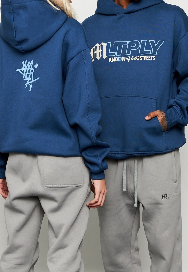 I felpe con cappuccio blu presentano grandi grafiche frontali e un logo più piccolo sul retro. Abbinati a pantaloni della tuta grigi chiari con tasche laterali.