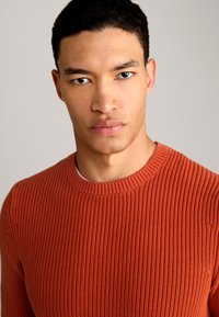 Maglione a girocollo lavorato a maglia in un ricco color terracotta, caratterizzato da un design a coste testurizzato e una vestibilità leggermente rilassata.