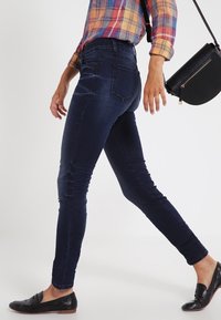 Mörkblå skinny jeans med slät textur, hög midja och bakfickor, tillsammans med svarta ballerinas och en liten axelväska.