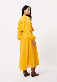 Robe jaune à manches longues avec un dos froncé, une taille cintrée et une jupe fluide. Associée à des bottes de cow-boy marron pour le contraste.