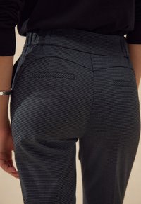 Pantaloni grigi a quadretti con vita alta e tasche laterali. Presentano un tessuto strutturato e una vestibilità sartoriale. Dettagli di cuciture visibili.