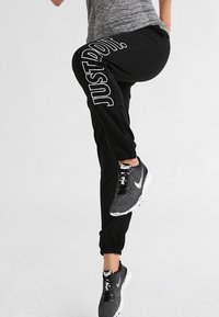 Persoon in zwarte trainingsbroek met de tekst "JUST DO IT.", grijze sportschoenen en een grijs topje, die één been optilt tegen een effen achtergrond.