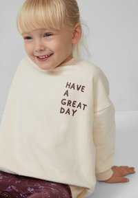 Kremowa bluza z prążkowanymi mankietami ozdobiona brązowym napisem "MIŁEGO DNIA". Miękki materiał, luźny krój, zestawiona z ciemnymi spodniami w wzory.