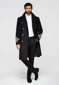 Schwarzer Wollmantel mit goldenen Knöpfen und bestickten Akzenten an den Ärmeln. Getragen über einem weißen Hemd und schwarzen Hosen, mit eleganten schwarzen Schuhen.