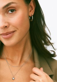 Hõbedased rõngas kõrvarõngad, kihilised hõbedased kaelakeed, millel on väike südame ripats, ja sile, neutraalse tooni bleiser.