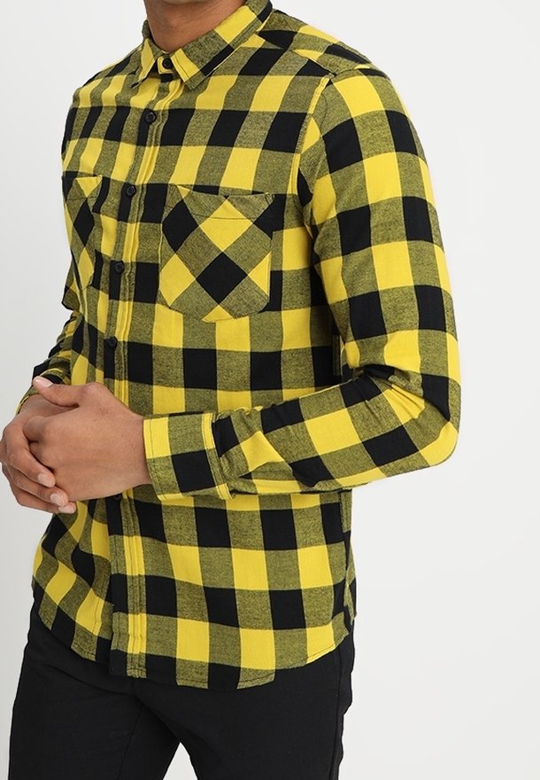 Homme portant une chemise à carreaux jaune et noir à manches longues avec poches poitrine boutonnées et pantalon noir, les mains jointes devant.