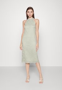 Vestido verde menta, com decote halter, sobreposição de renda apresentando padrões florais, comprimento até os meia-perna e silhueta reta. Usado com sandálias de tiras.