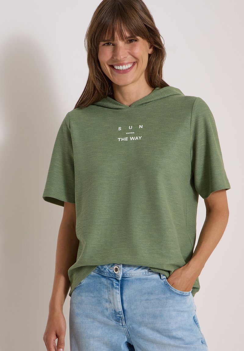 Femme souriante portant un t-shirt à capuche vert à manches courtes avec le texte "SUN KNOWS THE WAY" et un jean bleu clair, une main dans la poche.