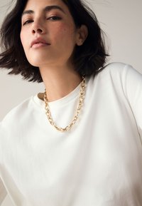White short-sleeve shirt with a rounded neckline, featuring a chunky gold chain necklace with oval links resting on the fabric.