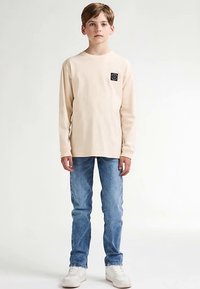 Světle béžové tričko s dlouhým rukávem a černou nášivkou s logem, v kombinaci s modrými džínami a bílými teniskami, stojící před jednobarevným pozadím.