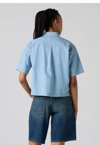 Camicia a maniche corte di colore blu chiaro con colletto, dotata di una vestibilità ampia e dettaglio della piega posteriore, abbinata a pantaloni corti in denim scuro.