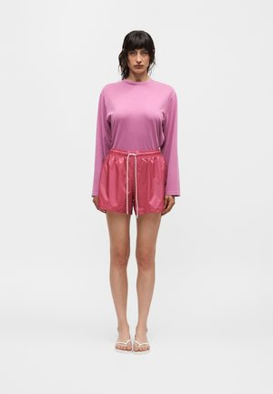 Femme debout portant un haut rose à manches longues, un short rose brillant avec cordon de serrage et des sandales blanches sur un fond blanc uni.