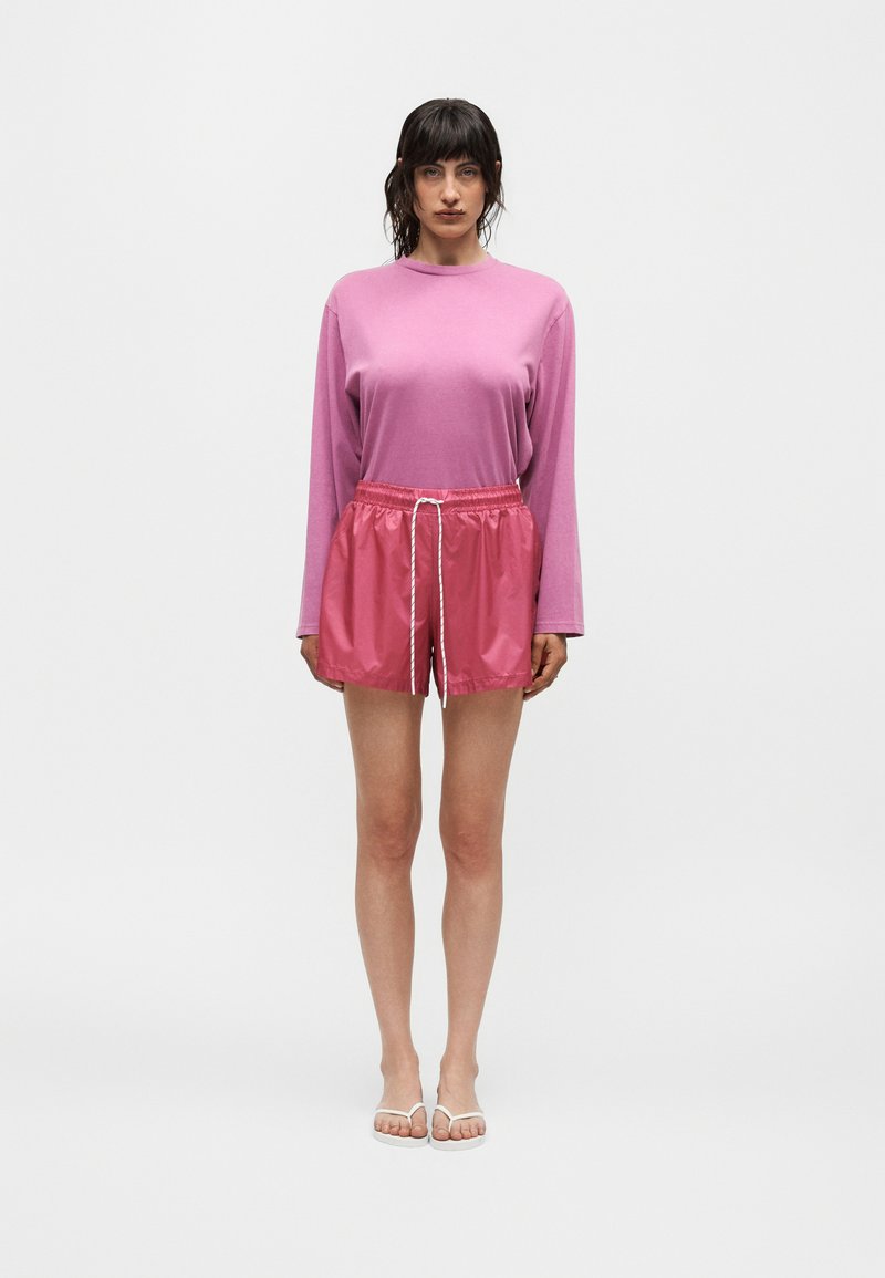 Femme debout portant un haut rose à manches longues, un short rose brillant avec cordon de serrage et des sandales blanches sur un fond blanc uni.