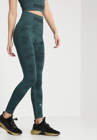 Kobieta ubrana w zielono-niebieskie legginsy w kamuflaż wzoru Puma oraz pasujący krótki top, czarne i złote sneakersy Puma, stojąca na tle białym.