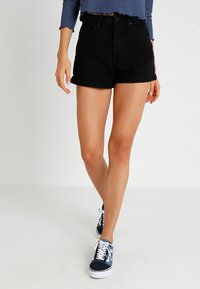 Femme portant un short en denim noir retroussé, un top court bleu et des baskets bleu et blanc à lacets, debout devant un fond blanc uni.