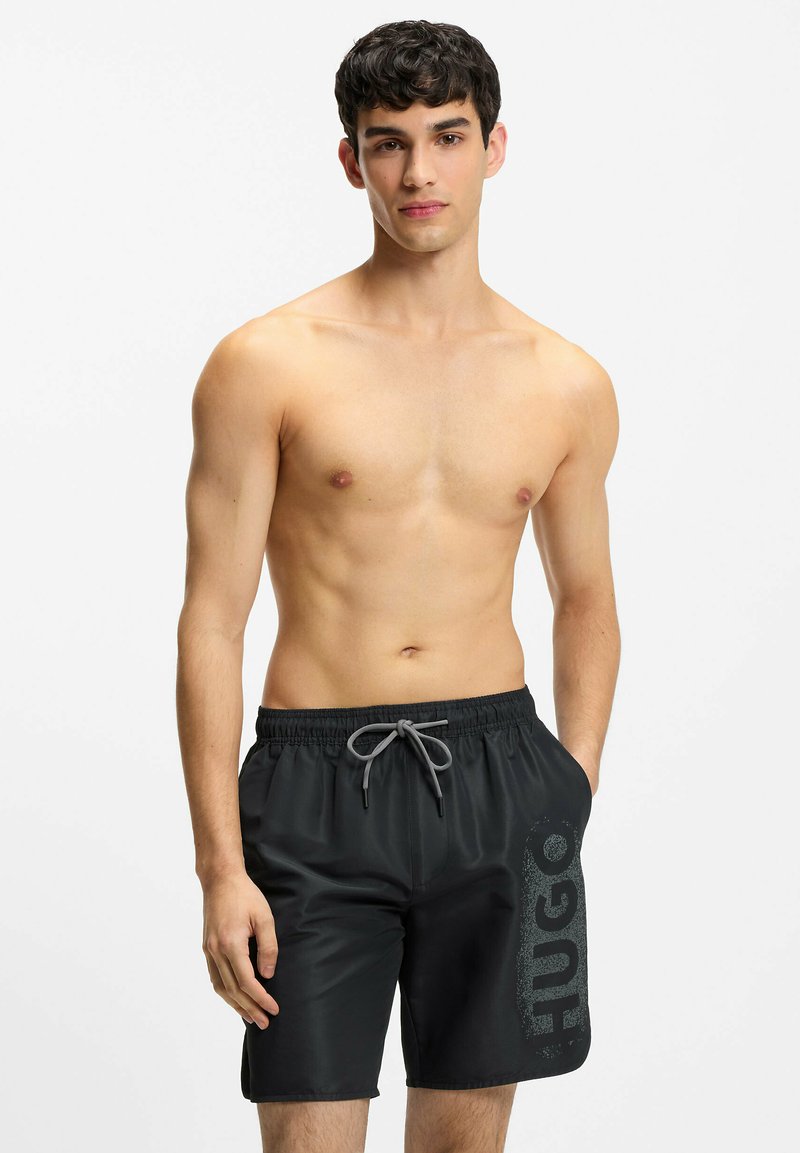 Jeune homme torse nu portant un short de bain noir avec un cordon gris et un grand logo « HUGO » sur la jambe gauche, les mains dans les poches.