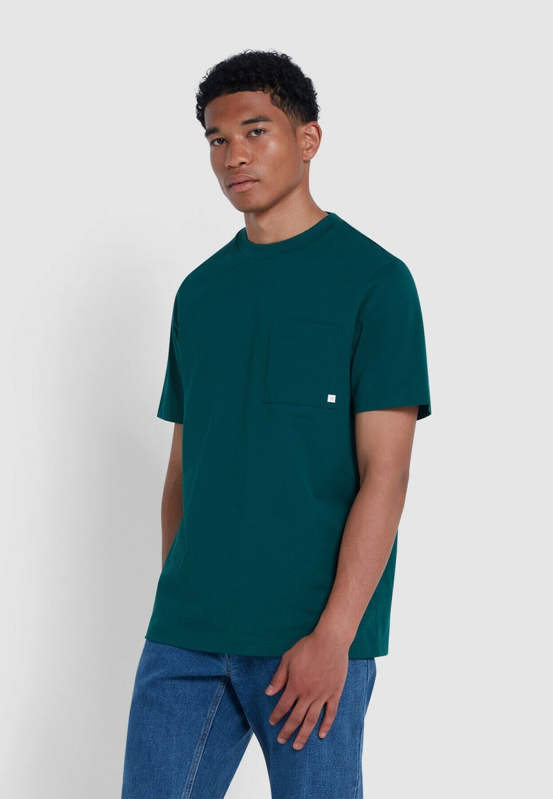 T-shirt a maniche corte color teal realizzato in cotone morbido, dotato di una tasca sul petto e collo rotondo. Abbinato a jeans in denim blu.