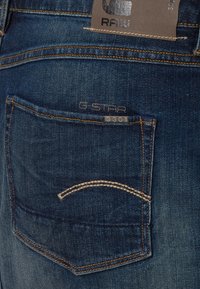 Mörkblå jeans i denim med en bakficka som har en böjd sömdesign, ett varumärkesmärke och subtila slitningsmönster.