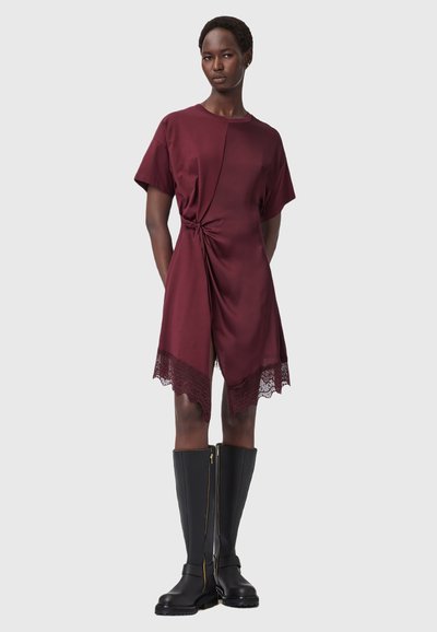 Robe bordeaux à manches courtes, avec un détail de nœud torsadé à la taille et une garniture en dentelle à l'ourlet. Bottes noires arrivee aux genoux avec une fermeture éclair dorée.