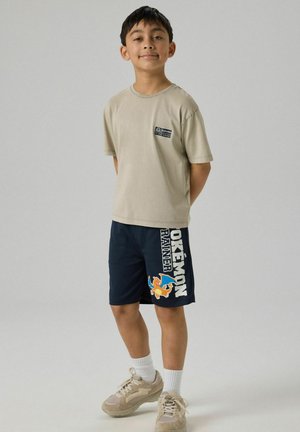 Jongen staat met handen achter de rug, draagt een beige T-shirt en marineblauwe Pokémon-short met Charizard-afbeelding.