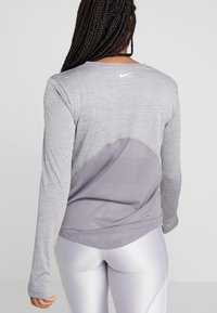 Kvinna med flätat hår som bär en grå långärmad träningsblus med nätbakpanel och vita figurnära leggings, stående mot en enfärgad bakgrund.