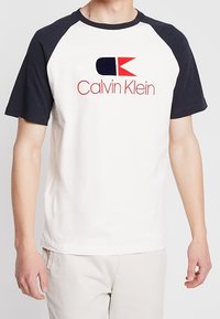 Camiseta de algodón blanca con mangas raglán en color marino, que presenta un logo en rojo y azul marino de "Calvin Klein" en el frente. Textura suave, corte clásico.