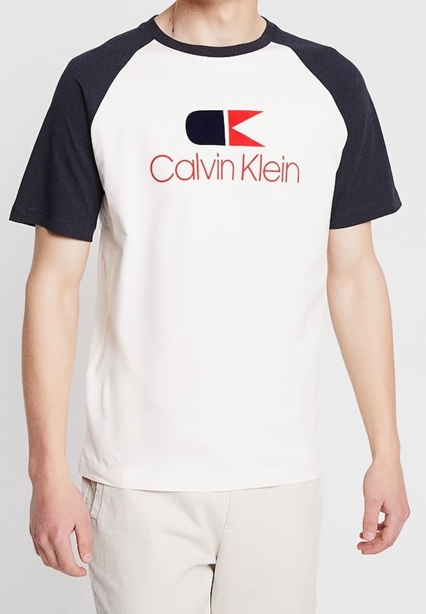 Camiseta de algodón blanca con mangas raglán en color marino, que presenta un logo en rojo y azul marino de "Calvin Klein" en el frente. Textura suave, corte clásico.